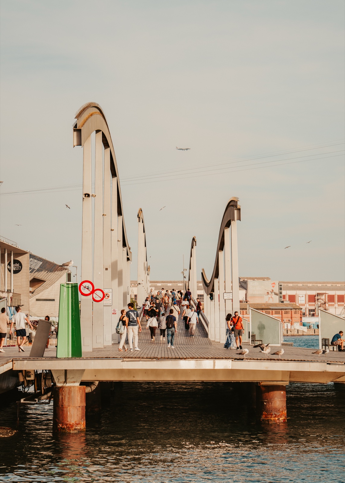 People crossing the Rambla de Mar footbridge between its curved arches, above the water